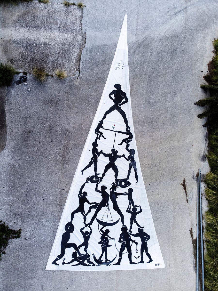 Aerial view of a triangular sail laid flat on concrete ground, painted with a black silhouette pyramid of human figures climbing and supporting one another.