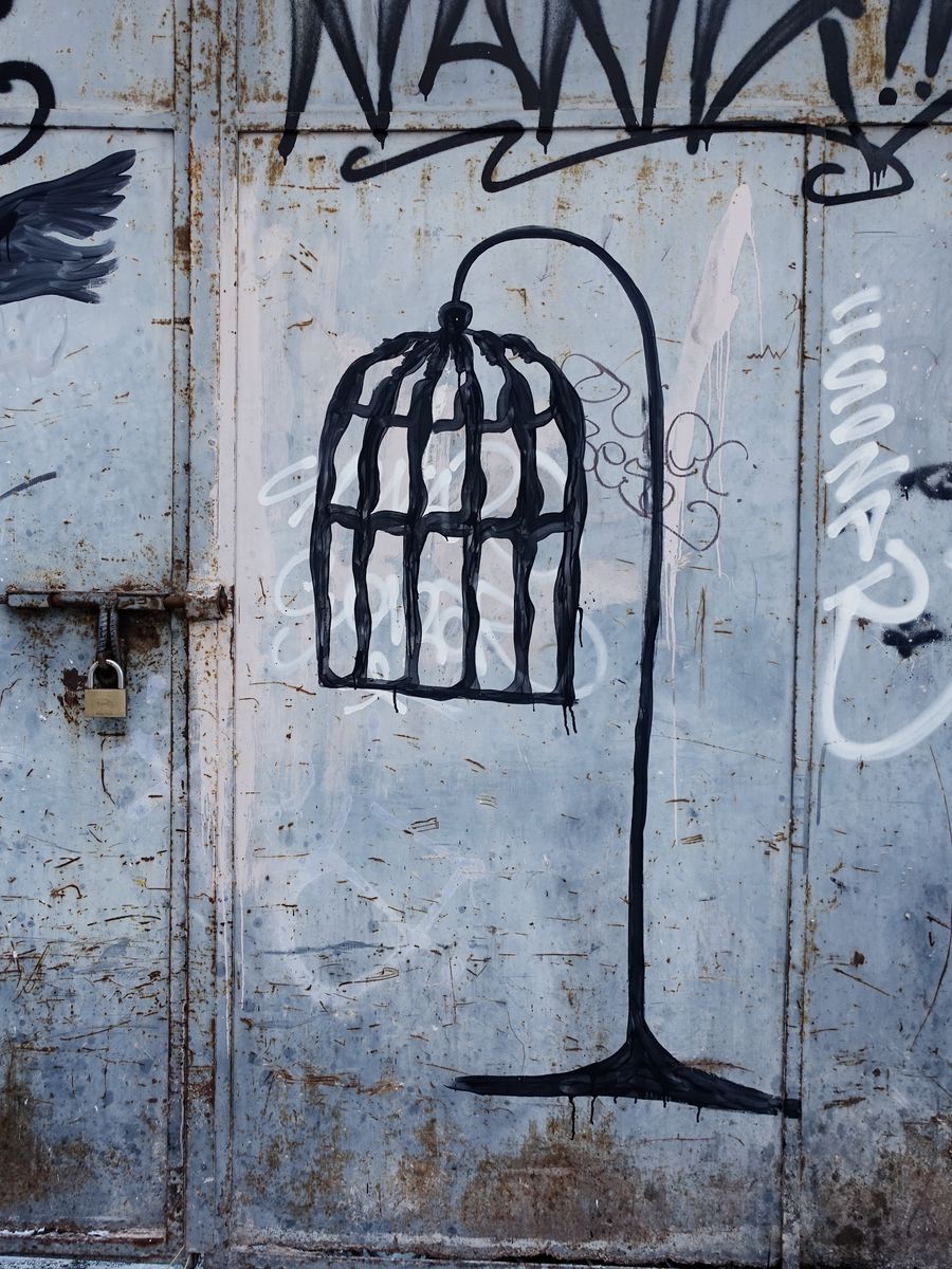 Black mural on a rusted metal door showing an empty bird cage hanging open from a curved stand, above a black wing painted separately on the left.