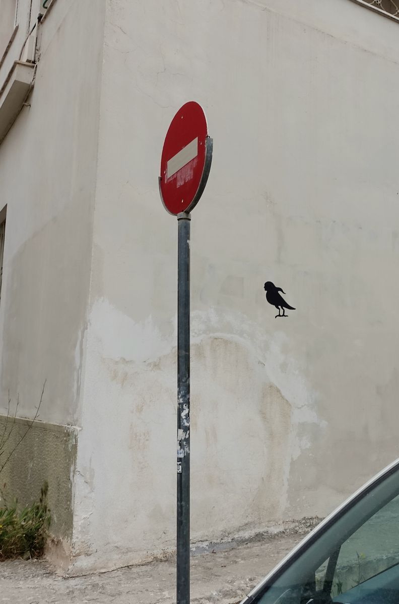 A no-entry traffic sign against a plain wall, with a small black bird silhouette painted on the wall beside it.