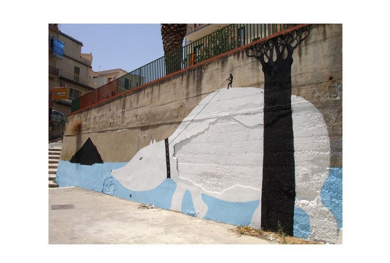 Large white silhouette of a polar bear painted on a concrete retaining wall beside a black baobab-like tree, a tiny figure walking along the top edge holding a rope, apartment buildings above. This section painted by Limow.