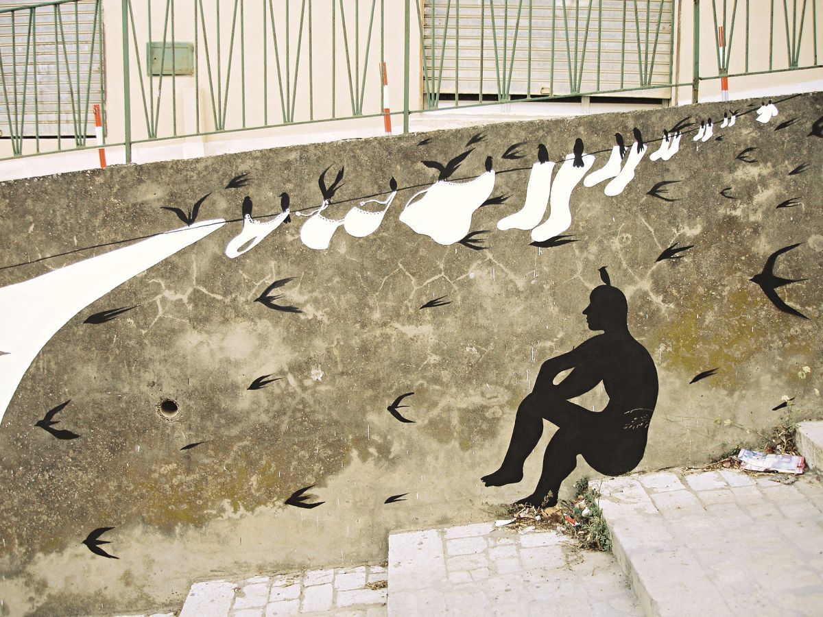 Black silhouette of a seated figure watching a row of black and white bird-shaped garments hanging from a clothesline strung along a concrete retaining wall, small swallows painted flying around them, steps leading up beside it.
