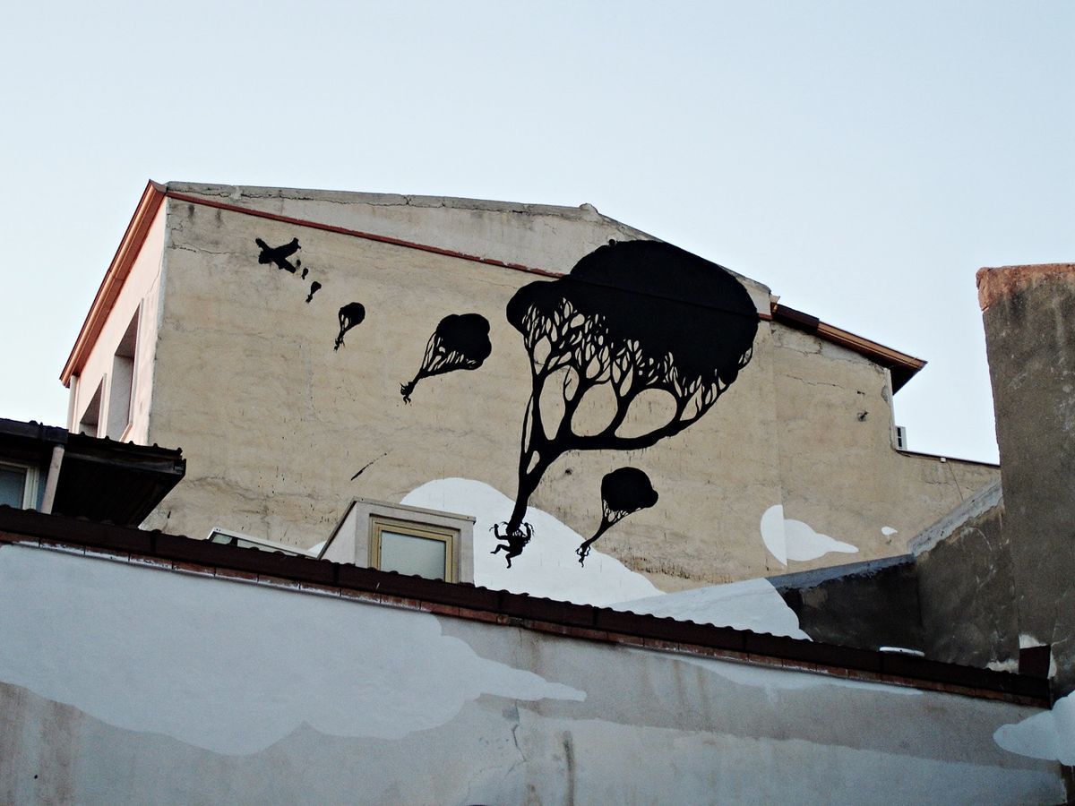 Black silhouette tree with a branching rounded canopy painted on a building gable beside a rooftop window, small parachuting figures and a crossed-out plane silhouette falling toward a white cloud shape below, seen against the sky above an adjoining roofline.