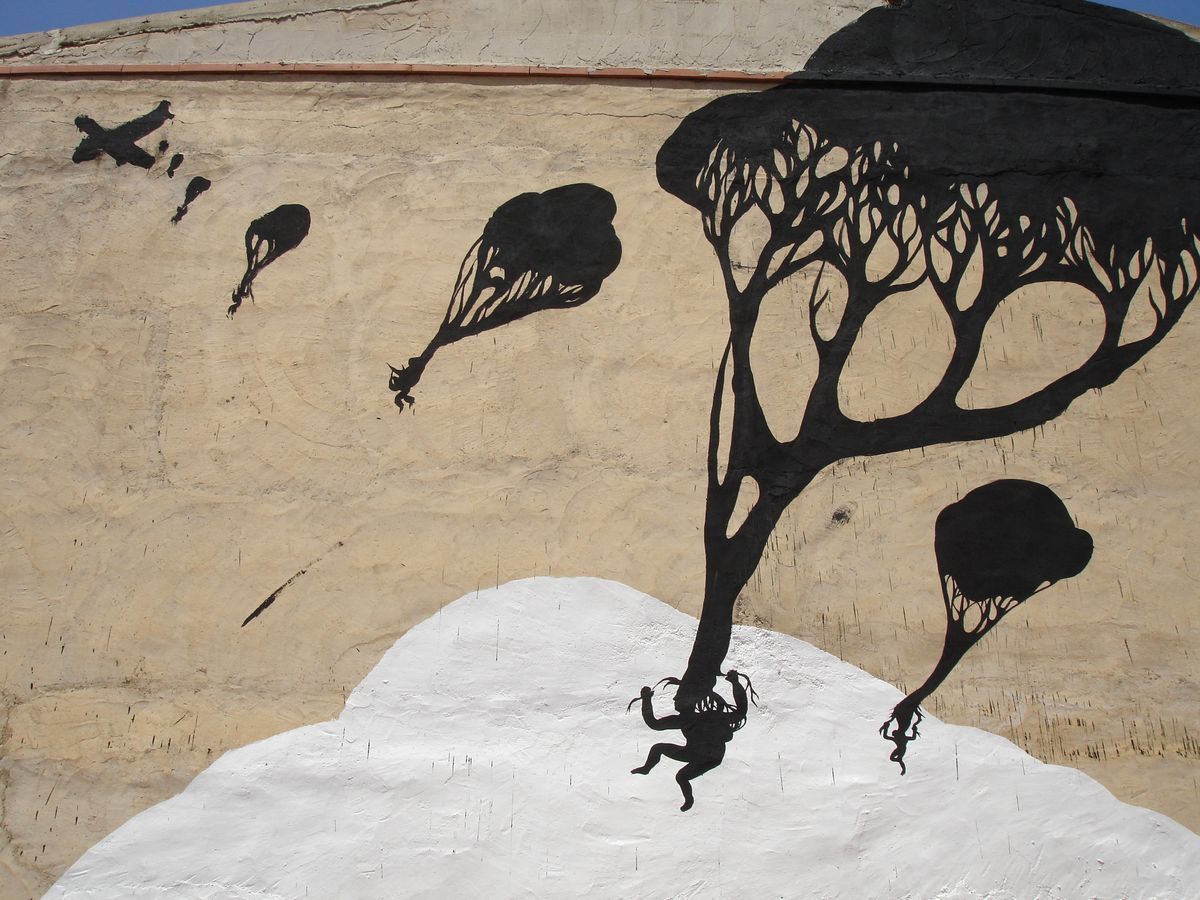 Black silhouette tree with a rounded canopy of branching leaf shapes painted on a tan building facade, small parachuting figures falling beside it toward a white cloud shape below, a crossed-out plane silhouette in the sky.