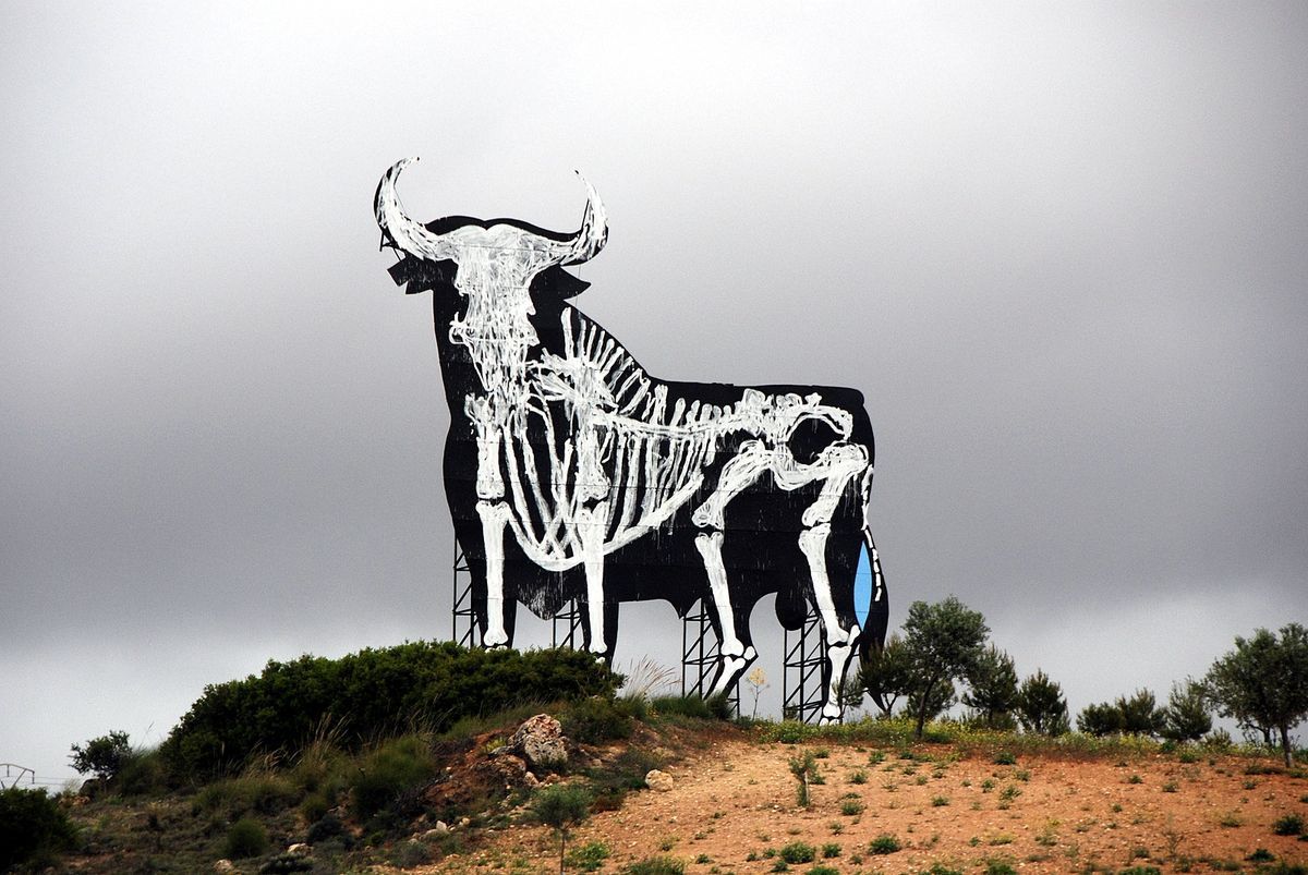 Large black Osborne bull billboard on a hillside with its skeleton painted in white over the silhouette, a small blue patch on its side, seen under an overcast sky.