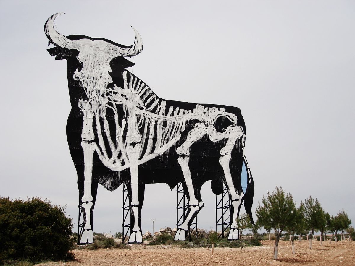 Close-up side view of the same black Osborne bull billboard with its full white skeleton painted across the silhouette, standing on a metal scaffold frame on a dirt hillside.