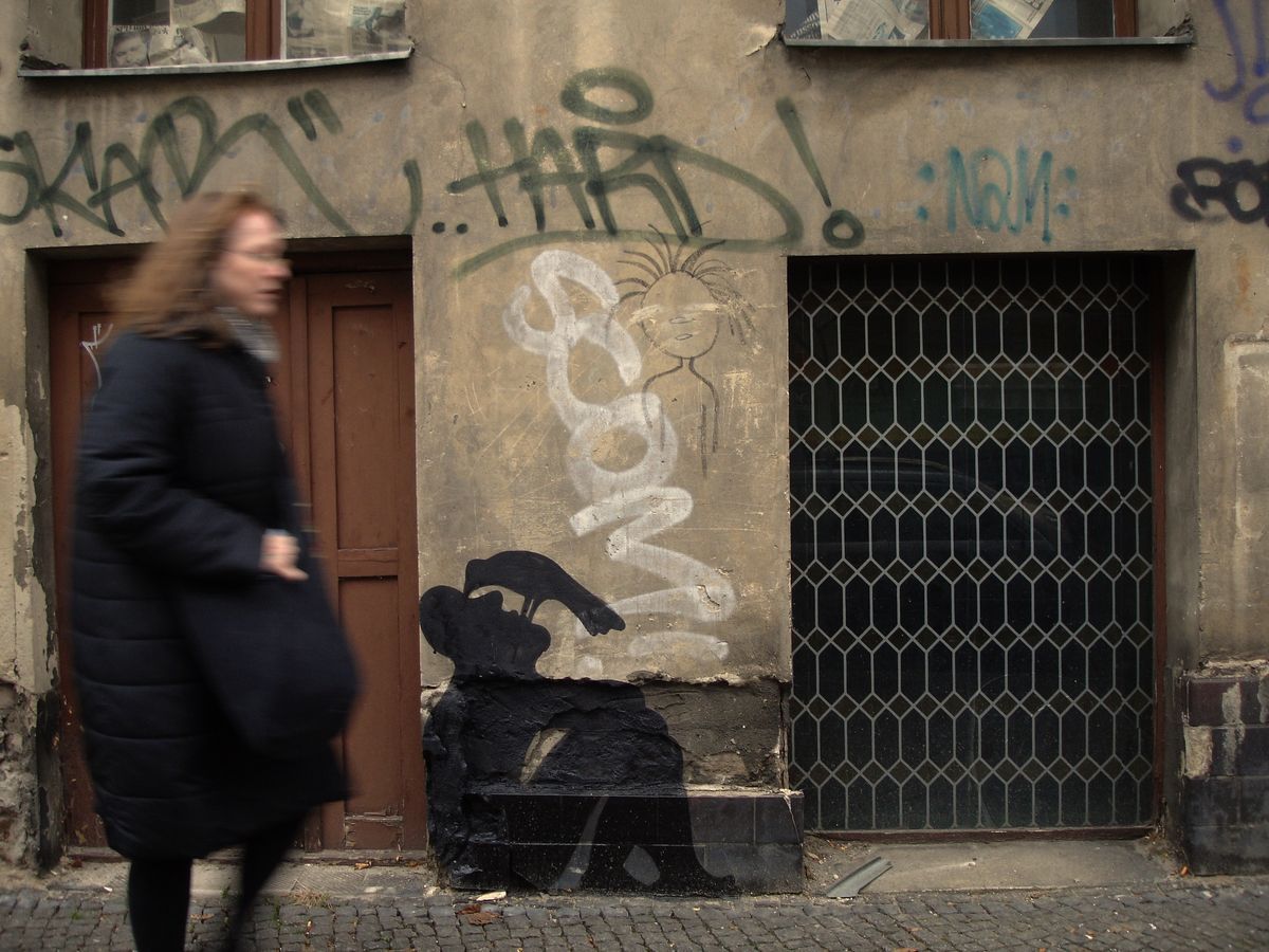 Small black silhouette of a seated figure with a bird perched beside its head, painted low on a weathered building base beside a wooden door, a woman walking past on the sidewalk, spray-painted graffiti and a chalk drawing above.