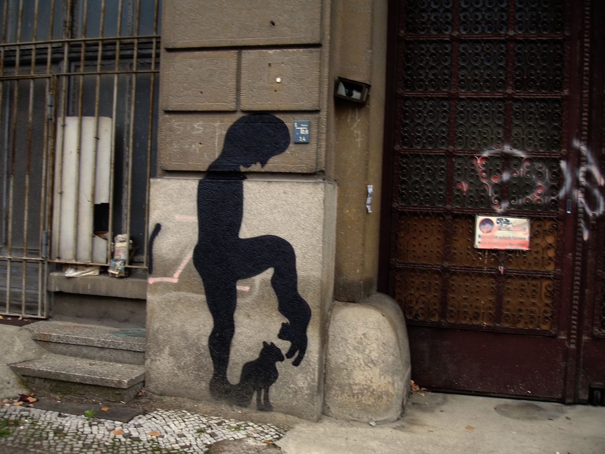 Black silhouette of a crouching nude figure balanced upside down on a stone plinth at a building corner, a small cat looking up at another cat dangling from the figure's hand, wrought iron gate and door beside it.