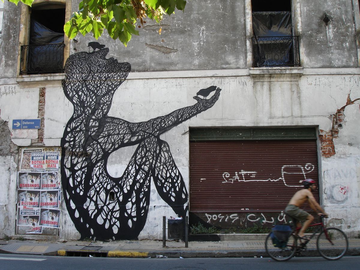 Black silhouette mural on a weathered building corner showing a crouching woman with a tree-root textured body reaching toward a small bird, pedestrians walking past below.