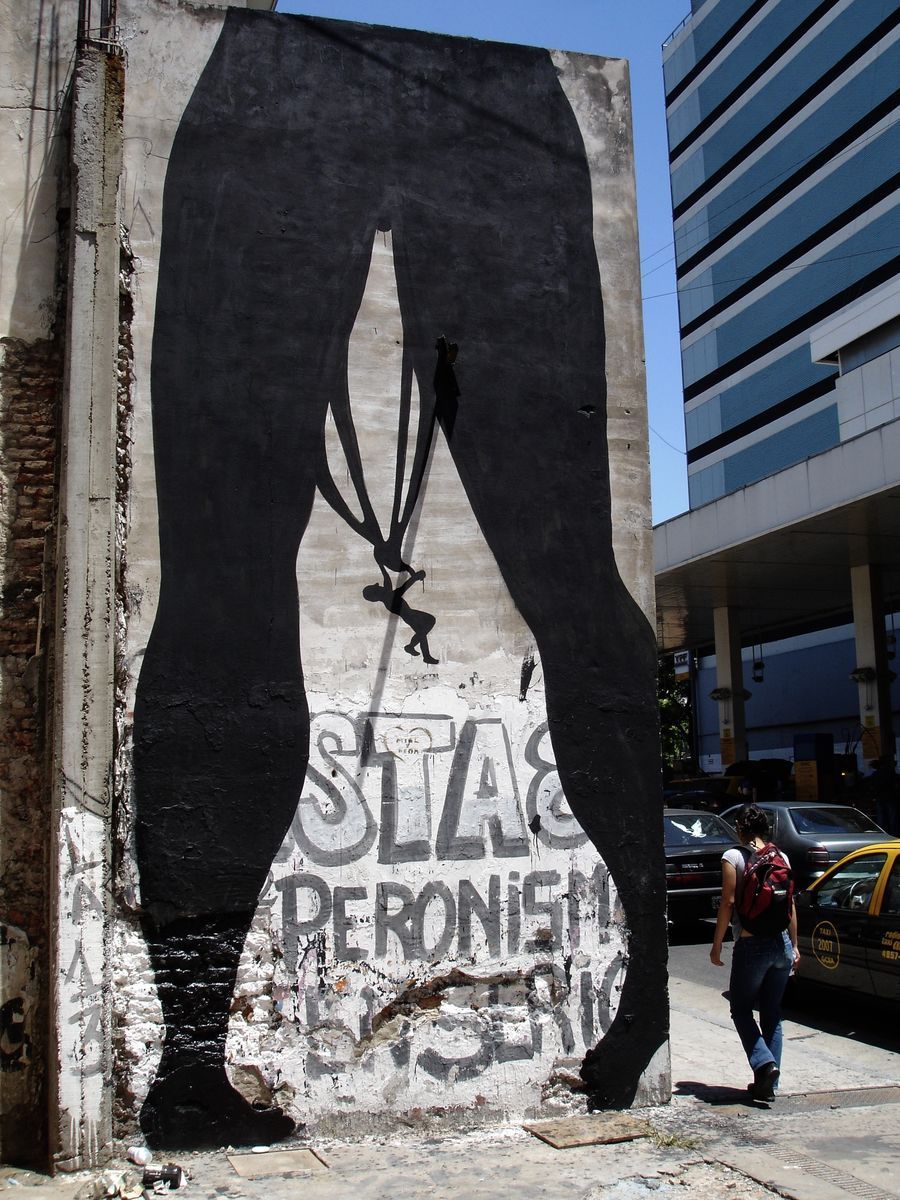 Tall black silhouette of a pair of legs painted on a building facade, a small figure hanging by its underwear from the crotch, faded political text below, a pedestrian and taxi passing on the street.