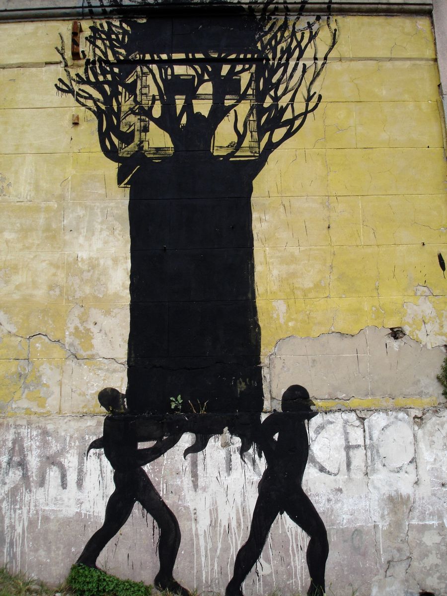 Black silhouette of a bare tree with a treehouse built into its branches, two standing figures below carrying the tree trunk horizontally between them, painted on a weathered yellow wall.