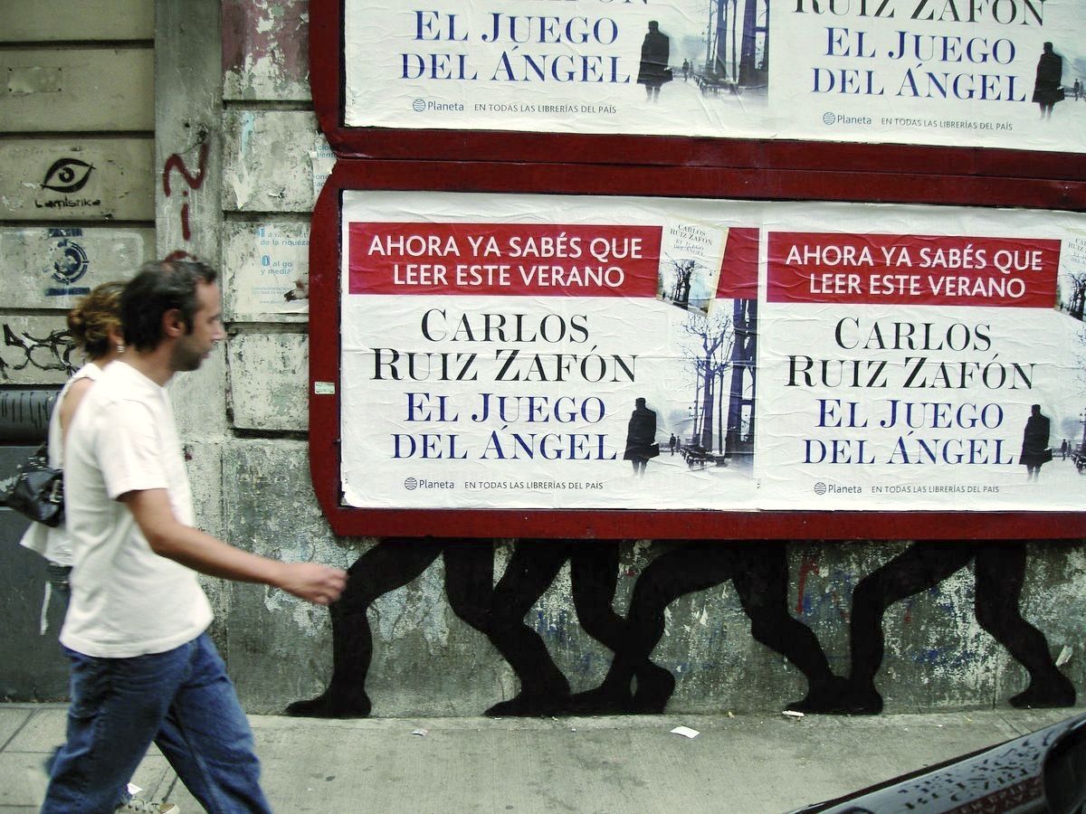 Row of black silhouette walking legs painted beneath a wall of layered book advertisement posters, a man in a white shirt walking past on the sidewalk.