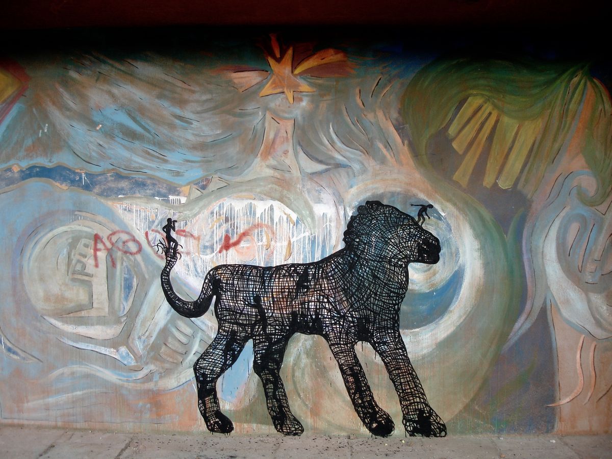 Black wireframe silhouette of a standing lion painted over a colorful abstract mural, a tiny figure perched on its tail and another on its back, on a wall beneath an underpass.