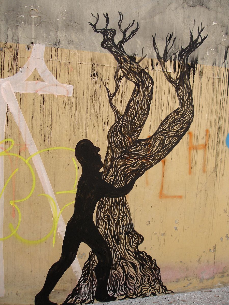 Black silhouette of a man embracing and lifting the trunk of an ornately patterned tree with bare branches, painted on a weathered plywood and concrete wall covered in graffiti.