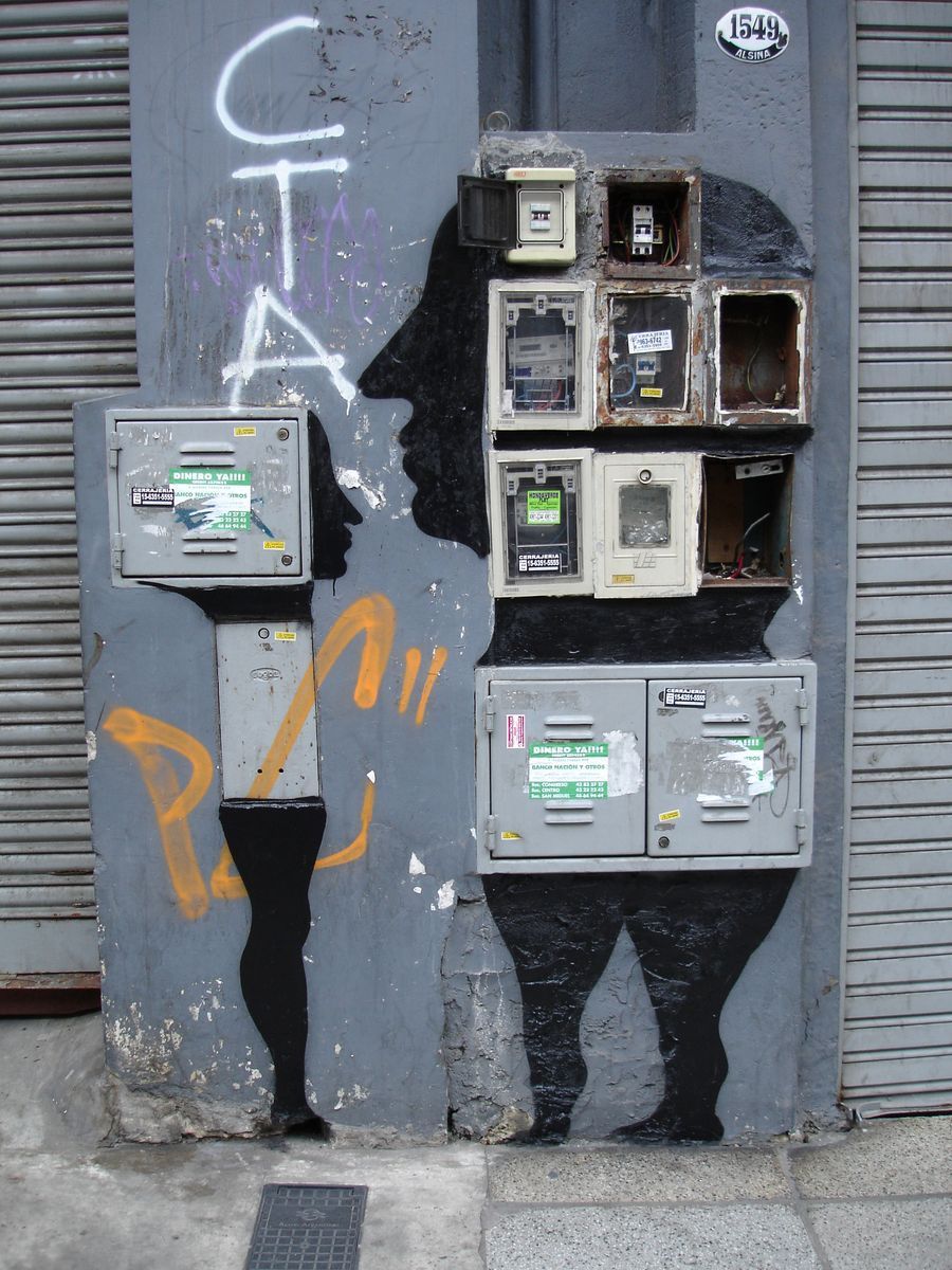 Black silhouette of a robot-like figure formed from a grid of electrical meter boxes set into a grey wall, graffiti tags scrawled beside it, a rolled metal shutter to the right.