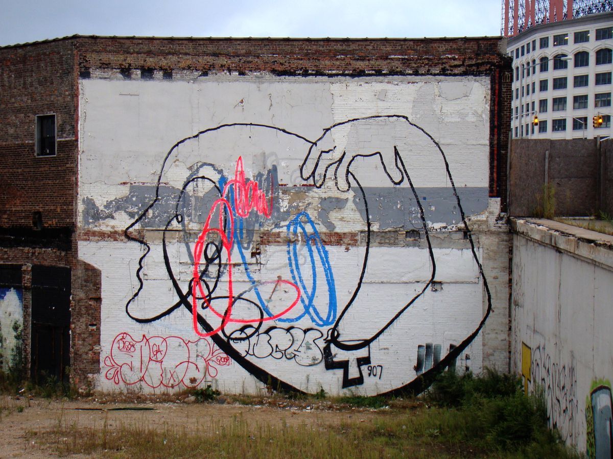 Black outline mural of a large profile head with a hand reaching from behind to touch its face, painted on a weathered brick and whitewashed warehouse wall, colorful graffiti scribbles inside the outline, city skyline visible behind.