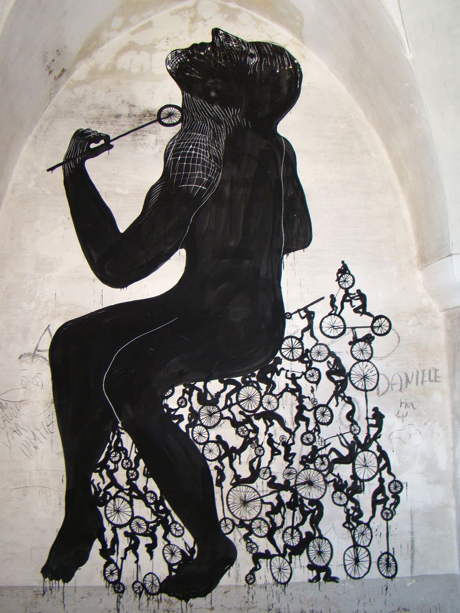Black silhouette of a giant seated figure holding a stick with a pinwheel, a swarm of tiny bicycles and riders climbing up its legs and torso like a tower, painted inside a vaulted stone archway.