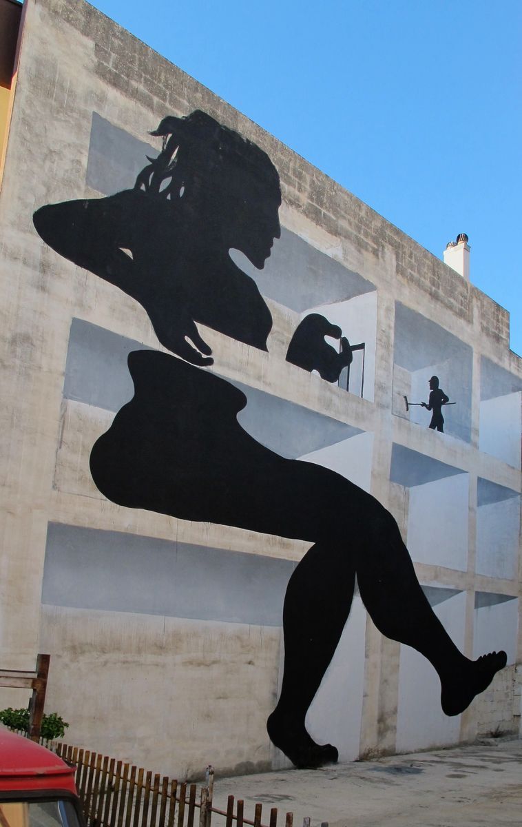 Building facade painted in trompe-l'oeil geometric panels with a black silhouette of a reclining nude woman stretched diagonally across the wall, small figures painted in some of the panels above her.