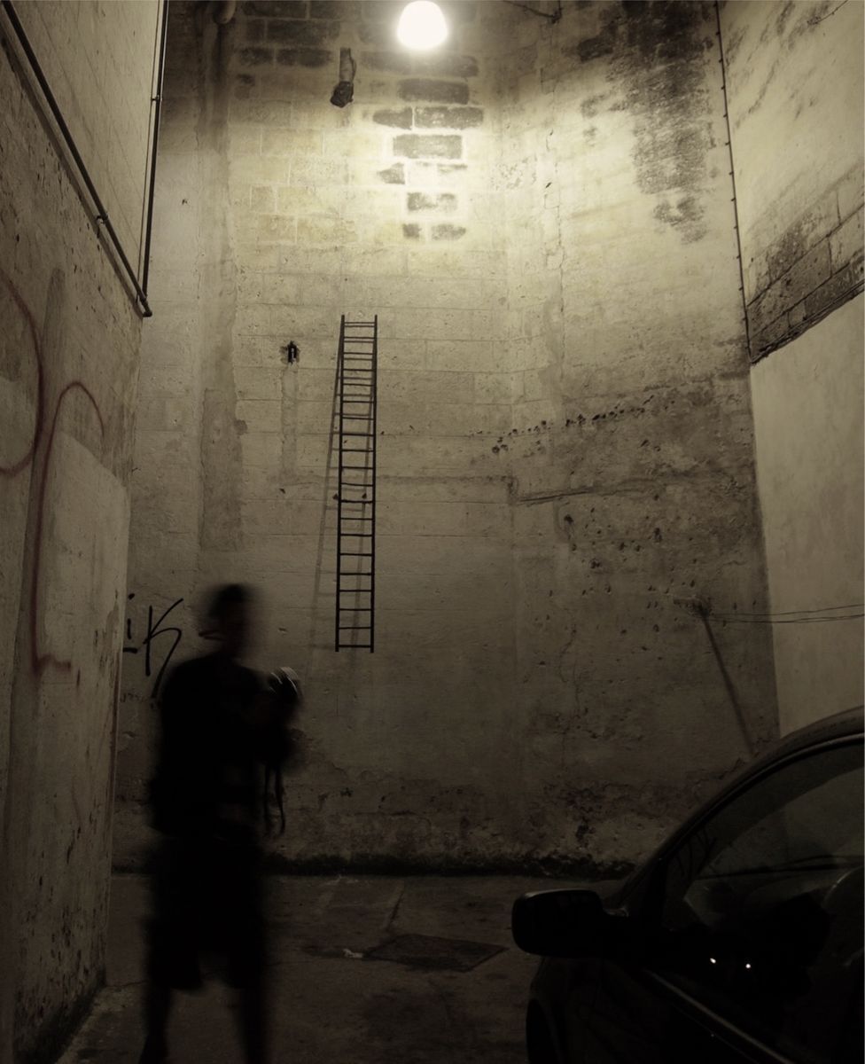 Tall black metal ladder leaning against a bare stone wall inside a dim vaulted room, its shadow cast beside it, graffiti marks on the wall to the left.